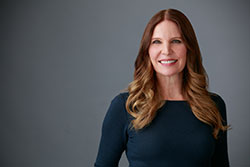 A woman with long hair stands confidently against a neutral background, smiling slightly towards the camera.