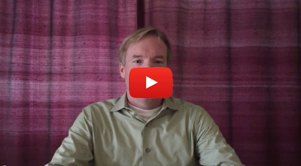 The image shows a man sitting at a desk with a red curtain behind him, wearing a light-colored shirt, and there is a YouTube icon overlaying the image.