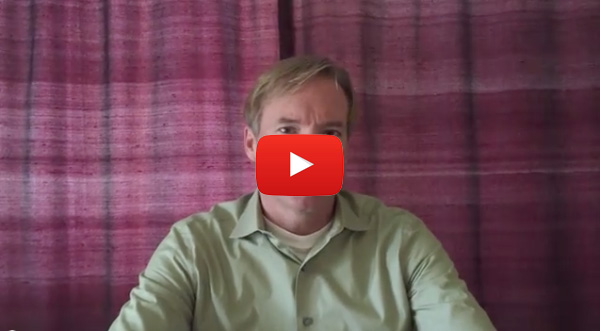 The image shows a man sitting at a table with a red background, wearing a light green shirt, and there s a YouTube logo overlaid on the screen.