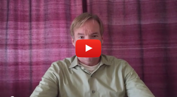 The image shows a man sitting at a table with a red background, wearing a light-colored shirt, looking directly into the camera with a neutral expression.