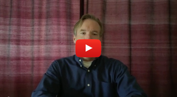 The image shows a man sitting in front of a red curtain with a YouTube video overlay on the screen.