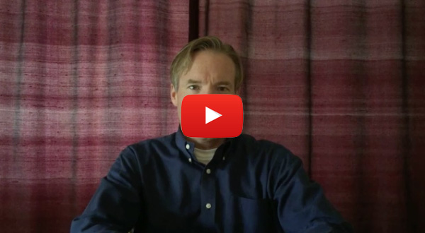 The image shows a man seated at a table with a laptop screen visible, looking directly at the camera with a serious expression. He has short hair and is wearing a button-up shirt. In the bottom right corner of the image, there s a YouTube icon with a play button, indicating that this is a screenshot from a video platform.