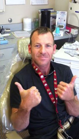 The image shows a man with a thumbs-up gesture, sitting in an office chair with his mouth open, wearing a lanyard with a name tag and a red shirt.