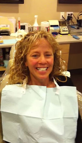 Woman seated in dental chair, smiling at camera, wearing blue mask over mouth.