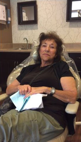 The image shows an elderly woman sitting in a dental chair with protective eyewear, looking down at a piece of paper, likely in a dental office setting.