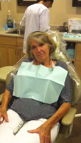 An elderly woman wearing a dental mask while seated in a dental chair with a hygienist standing behind her.