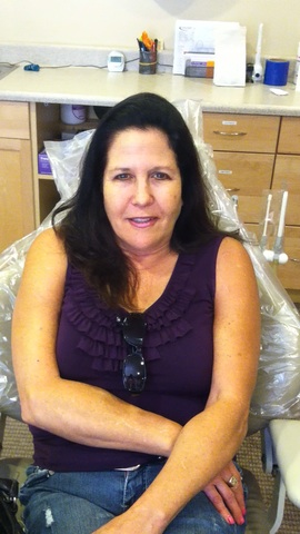 The image shows a woman sitting in a dental chair with her legs crossed, wearing a purple top and sunglasses on her head, looking directly at the camera with a slight smile.