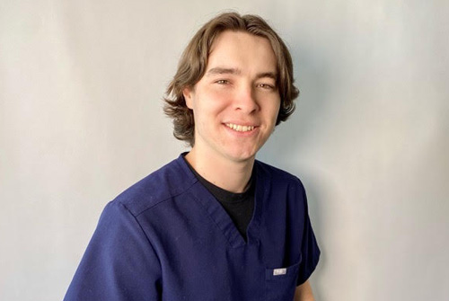 The image shows a man wearing scrubs, smiling at the camera.
