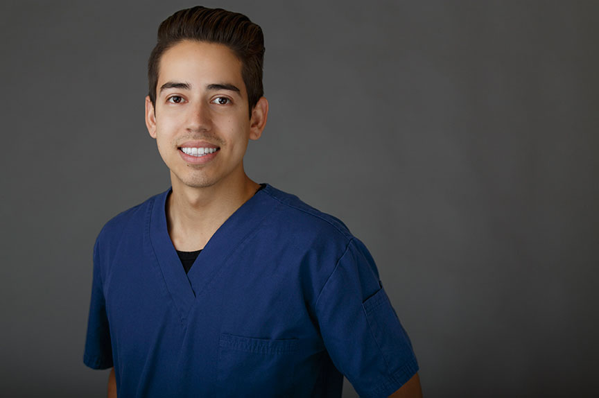 A man wearing scrubs stands against a dark background with a neutral expression.