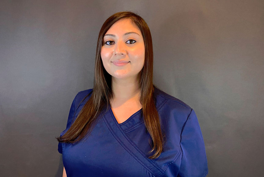 A woman wearing a blue scrub top stands against a dark background.