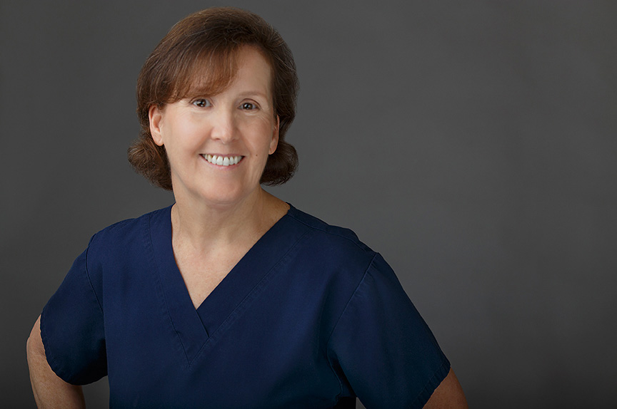 Woman in blue scrubs standing against dark background.