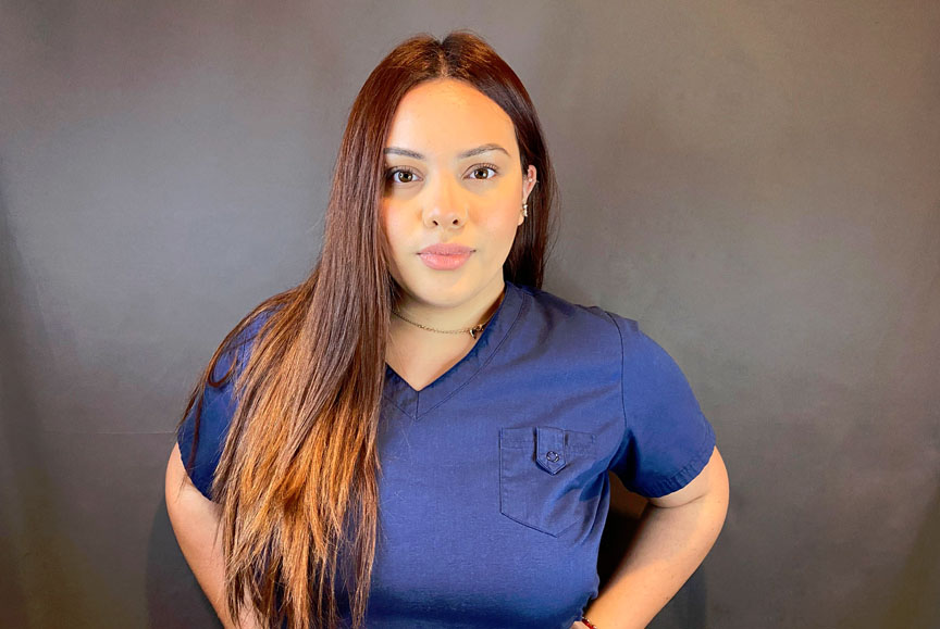 The image shows a woman standing against a dark background, wearing a blue shirt and white pants, with her hair styled down.