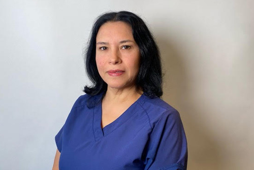 The image shows a woman wearing a blue scrub top, standing against a white background with a neutral expression on her face.