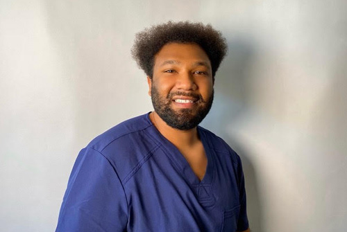 The image shows a man wearing scrubs and smiling at the camera.