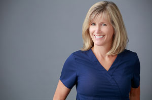 A woman wearing a blue shirt stands against a white background, smiling slightly at the camera.