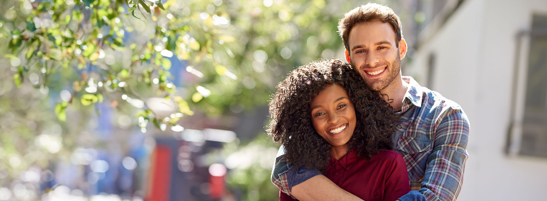 A man and woman are hugging each other outdoors  the man is wearing a plaid shirt and has short hair, while the woman is wearing a dark top with a lighter sleeve and has long hair.