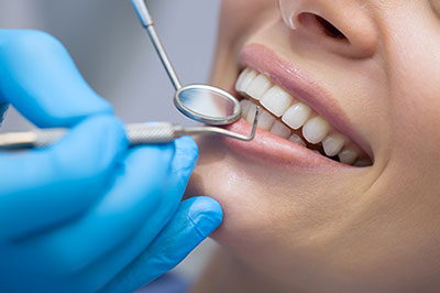 A woman receiving dental care with a smiling expression, while a dental hygienist works on her teeth using tools visible through a magnifying glass.
