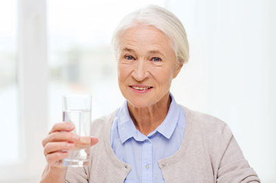 The image shows an elderly woman holding a glass of water with her right hand while standing indoors.