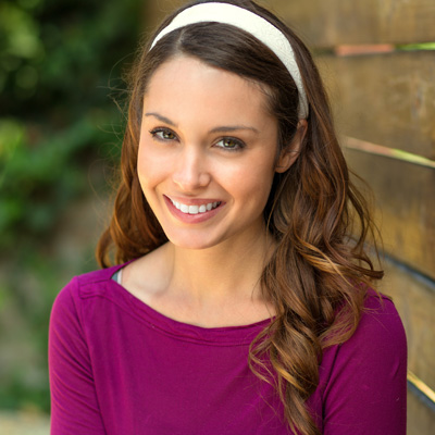 A young woman with long hair is smiling at the camera, wearing a pink top and headband, standing against a wooden fence.